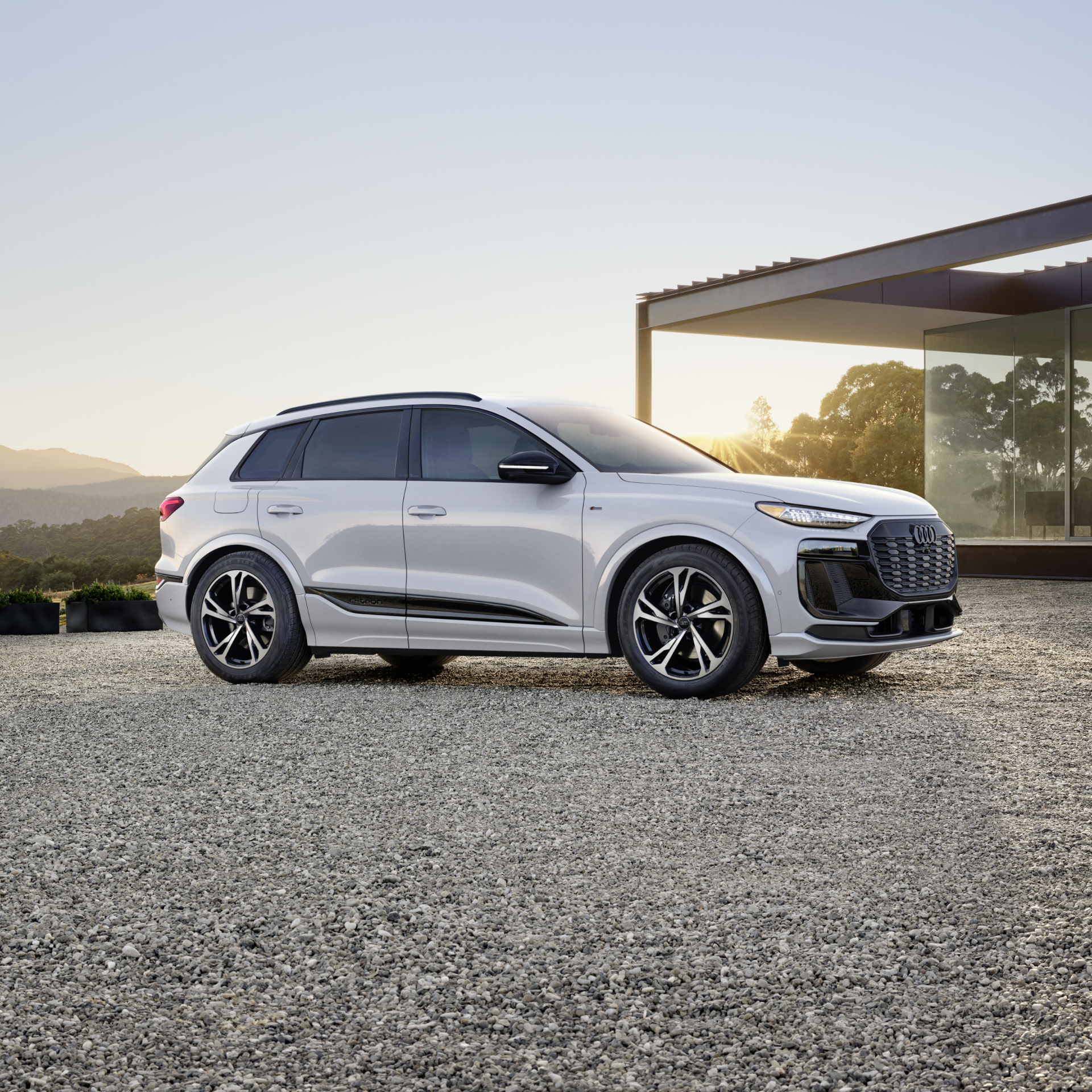 A white Audi Q6 e-tron electric SUV parked on a gravel driveway in front of a modern glass house at sunrise.